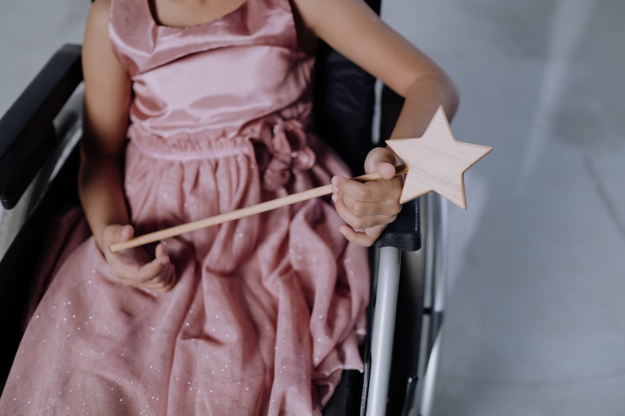 A child in a pink dress holding a star-shaped magic wand, seated in a wheelchair.